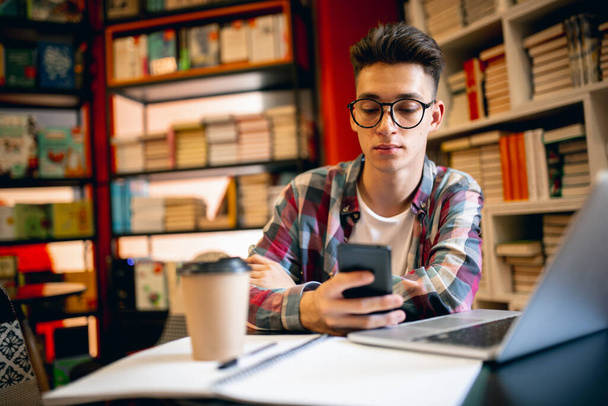 Een jonge blanke man, student zit aan tafel met laptop over huis of bibliotheek achtergrond. - Foto, afbeelding