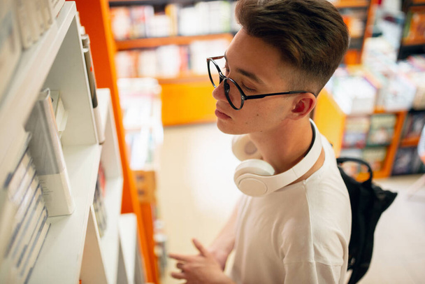 The male college student with book standing near shelf in library. - Foto, Imagen