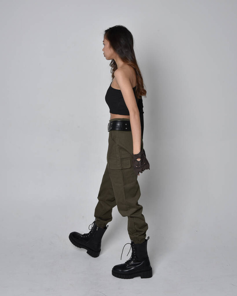 Full length portrait of pretty brunette, asian girl wearing black top and khaki utilitarian army pants and leather boots. Standing pose with gestural hands, walking away from the camera.  isolated against a light grey studio background. - Фото, изображение