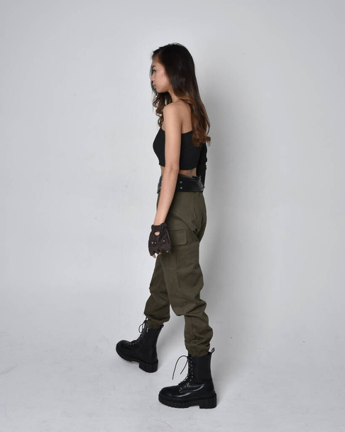 Full length portrait of pretty brunette, asian girl wearing black top and khaki utilitarian army pants and leather boots. Standing pose with gestural hands, walking away from the camera.  isolated against a light grey studio background. - Foto, Imagen
