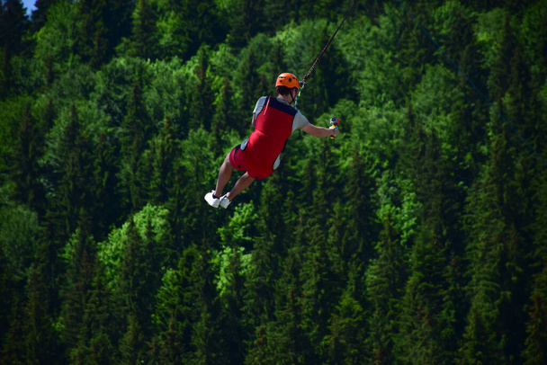 Młody człowiek w kasku wyskoczył z bungee jumping i teraz wisi na linie, huśtając się i filmując się na sportowej kamerze wideo na niewyraźnym tle zielonego lasu - Zdjęcie, obraz