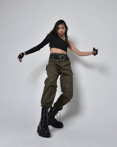 Uzun boylu, esmer, siyah bluz giyen Asyalı bir kız ve asker pantolonu ve deri çizmeleri. Açık gri bir stüdyo arka planında, el hareketleriyle poz veriyor.. - Fotoğraf, Görsel
