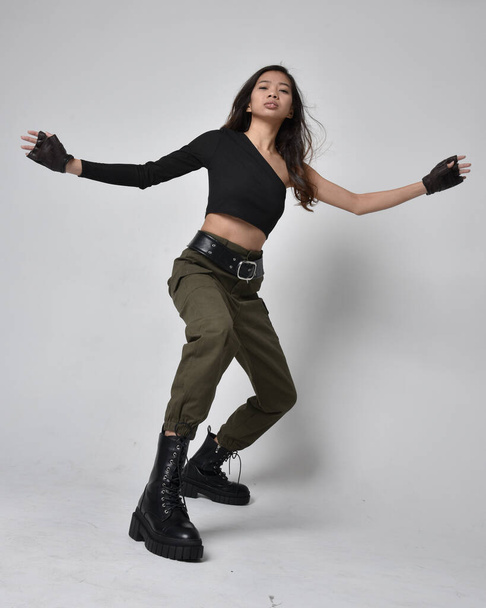Full length portrait of pretty brunette, asian girl wearing black top and khaki utilitarian army pants and leather boots. Standing pose with gestural hands, isolated against a light grey studio background. - Foto, Imagen