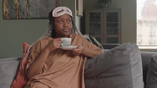 Medium shot van jonge pensive vrouw van Afrikaanse etniciteit het dragen van huiskleding en slaapmasker op voorhoofd het drinken van thee tijdens het zitten op de bank in gezellige woonkamer - Video