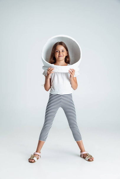 Full-length portrait of of beautiful preschool girl in big white astronaut helmet isolated on gray studio background. Childhood, education, emotion concept - Фото, изображение