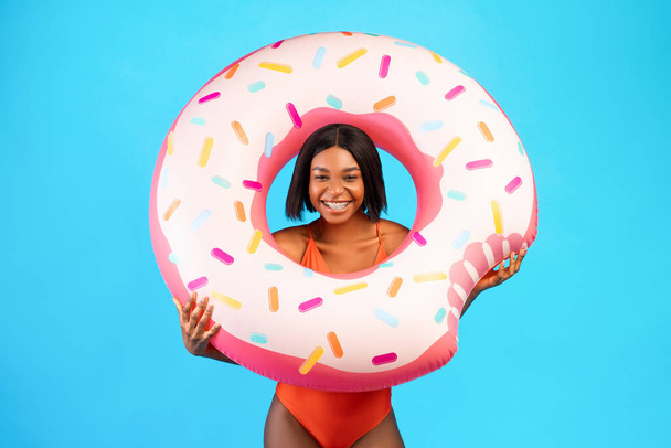 Mayo giymiş, donut şeklinde şişirilebilir bir yüzükle poz veren, mavi stüdyo arka planında kameraya gülümseyen genç siyahi bir bayan. - Fotoğraf, Görsel