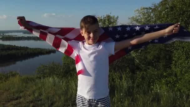 Menino acenando bandeira nacional dos EUA ao ar livre sobre o céu azul na margem do rio - Filmagem, Vídeo