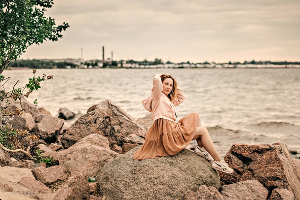 若い女性が海岸の岩の上に座って夕方の海を見て. - 写真・画像