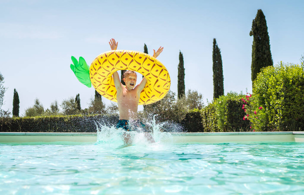 Cheerful Screaming boy in yellow pineapple inflatable ring having fun and jumping into swimming pool. Merry childhood or summer holidays concept image. - Foto, afbeelding