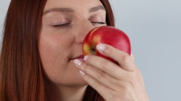 Onnellinen nuori nainen pitää tuoretta kypsää punaista omenaa kädessä ja haisee orgaanisille hedelmille, jotka on eristetty harmaalla pohjalla. Terveellinen syöminen. - Materiaali, video