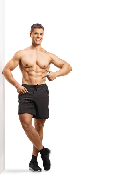 Full length portrait of a smiling shirtless man wearing shorts and pointing at his abs isolated on white background - Fotoğraf, Görsel