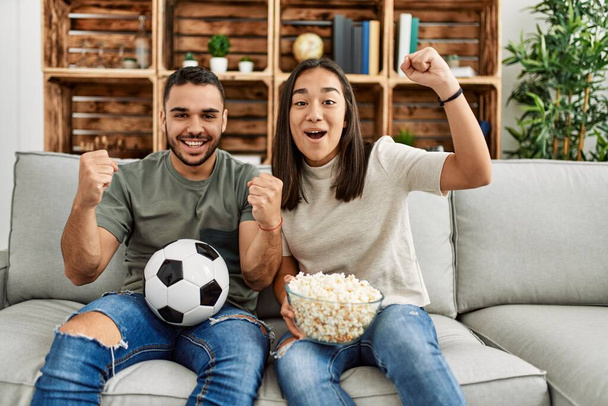 Νεαρό ζευγάρι Λατίνων βλέποντας ποδόσφαιρο αγώνα τρώει καλαμπόκι στο σπίτι. - Φωτογραφία, εικόνα