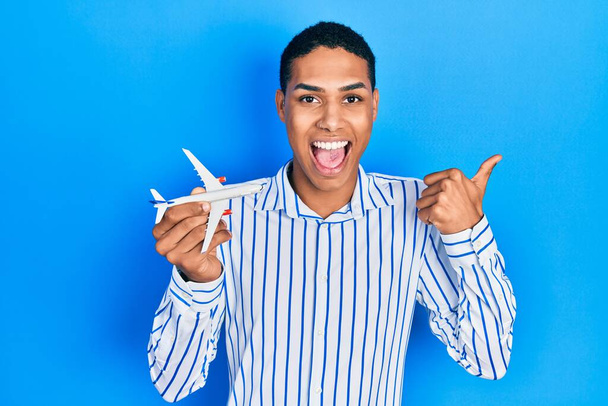Mladý africký Američan chlap drží letadlo hračka ukazující palec nahoru na stranu s úsměvem šťastný s otevřenými ústy  - Fotografie, Obrázek
