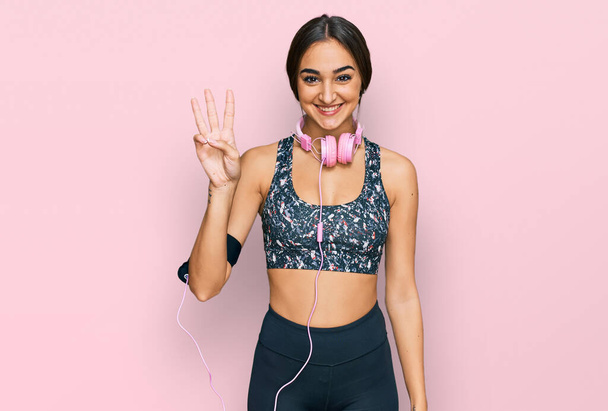 Beautiful brunette woman wearing gym clothes and using headphones showing and pointing up with fingers number three while smiling confident and happy.  - Photo, Image