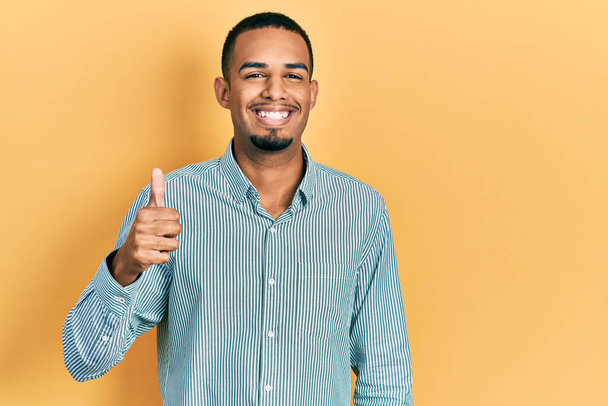 Joven afroamericano hombre vistiendo ropa casual sonriendo feliz y positivo, pulgar hacia arriba haciendo excelente y signo de aprobación  - Foto, Imagen