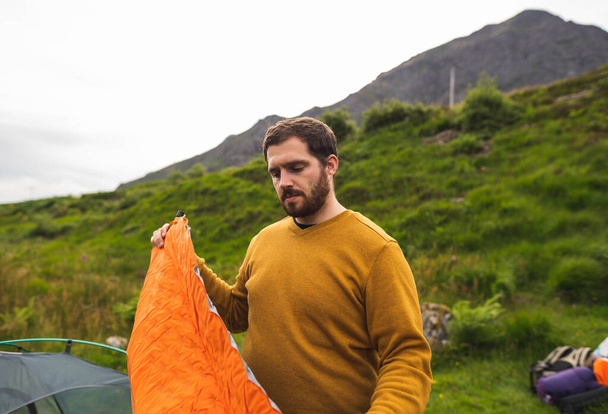 Молодой и привлекательный мужчина, раздувающий матрас рядом со своей палаткой для самостоятельного похода в Шотландию. Оборудование для путешествий во время отпуска - Фото, изображение