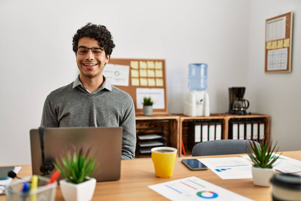 Junger hispanischer Mann im Business-Stil sitzt am Schreibtisch im Büro und blickt mit sexy Gesichtsausdruck, fröhlichem und glücklichem Gesicht in die Kamera.  - Foto, Bild