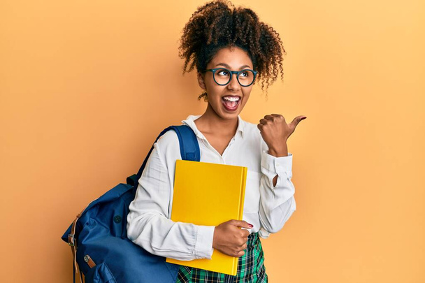 Belle femme afro-américaine aux cheveux afro portant un sac d'école et tenant des livres pointant le pouce vers le côté souriant heureux avec la bouche ouverte  - Photo, image