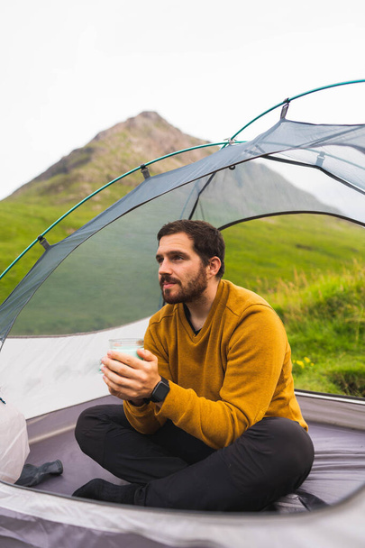 Young and attractive man sitting inside a tent outdoors enjoying a hot beverage in a reusable cup in the middle of nature in Scotland - Foto, Imagem