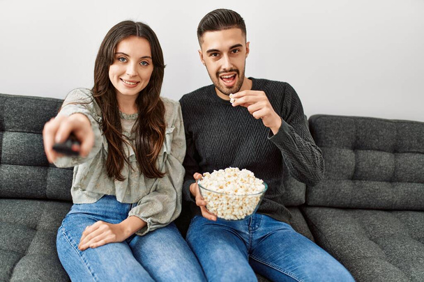 Fiatal spanyol házaspár filmet néz és popcornt eszik otthon a kanapén.. - Fotó, kép