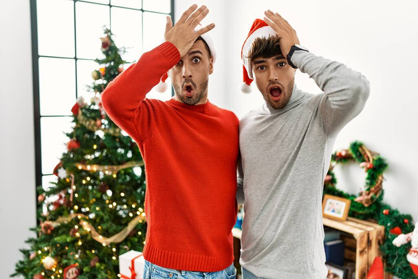Young gay couple standing by christmas tree wearing hat surprised with hand on head for mistake, remember error. forgot, bad memory concept.  - Photo, Image
