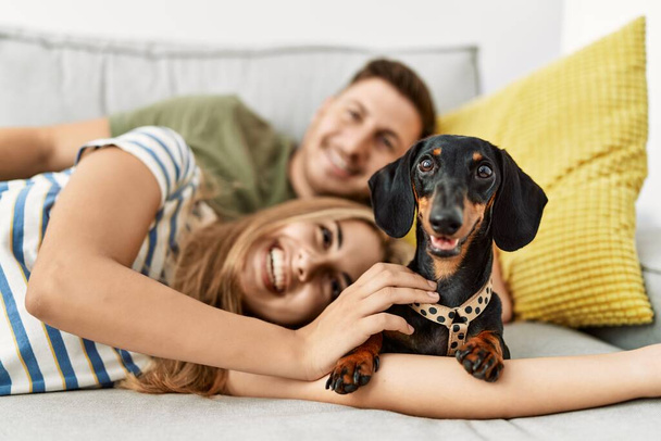 Νεαρό ζευγάρι ισπανόφωνων χαμογελαστό χαρούμενο ξαπλωμένο στον καναπέ με σκύλο στο σπίτι. - Φωτογραφία, εικόνα