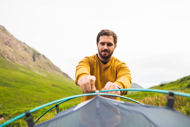 Foto de um jovem e atraente homem montando uma tenda de acampamento ao ar livre para desfrutar de um belo dia no meio da natureza na Escócia.  - Foto, Imagem