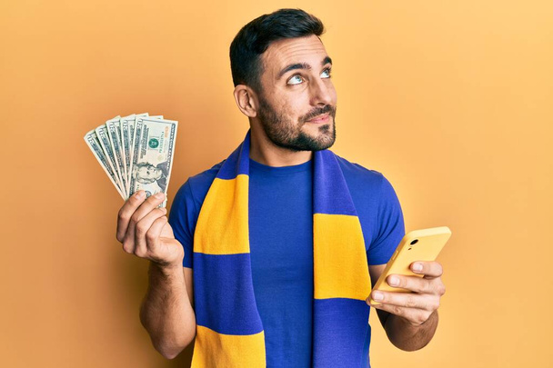 Jonge Spaanse man voetbal supporter met behulp van smartphone met Amerikaanse dollars bankbiljetten glimlachend kijken naar de zijkant en staren weg denken.  - Foto, afbeelding