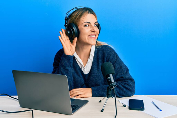 Giovane donna caucasica che lavora allo studio radiofonico sorridendo con mano sull'orecchio ascoltando voci o pettegolezzi. concetto di sordità.  - Foto, immagini