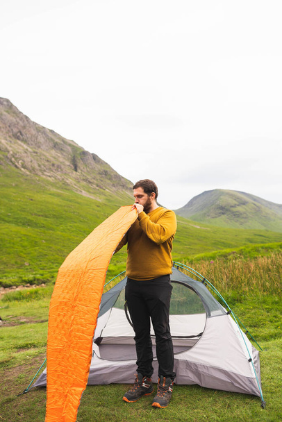 Joven y atractivo hombre inflando un colchón al lado de su tienda de campaña para hacer camping en solitario en Escocia. Equipamiento para viajar durante las vacaciones - Foto, Imagen