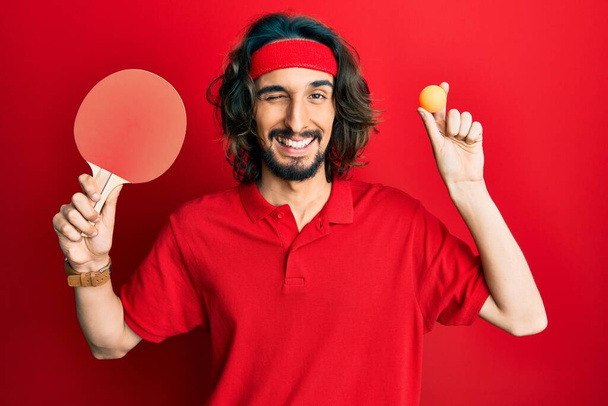 Mladý hispánec drží červený ping pong raketa a míček mrkání na kameru se sexy výraz, veselý a šťastný obličej.  - Fotografie, Obrázek