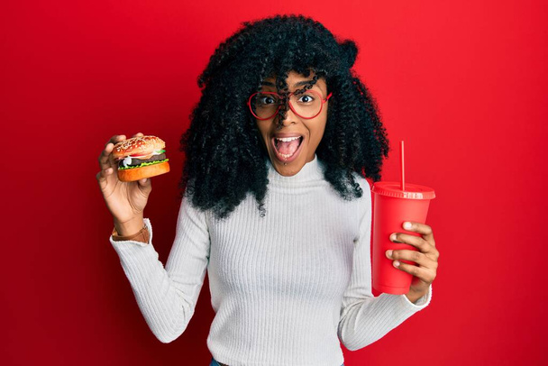 Afroamerikanerin mit Afro-Haaren isst einen leckeren klassischen Burger und Limo feiert verrückt und erstaunt über den Erfolg mit offenen Augen schreit aufgeregt.  - Foto, Bild