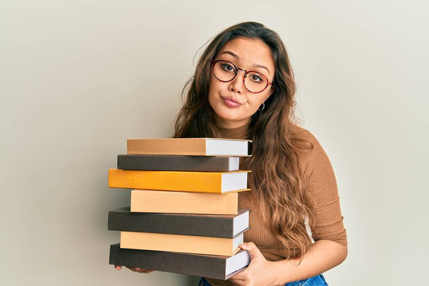 Young hispanic girl holding a pile of books skeptic and nervous, frowning upset because of problem. negative person.  - Photo, Image