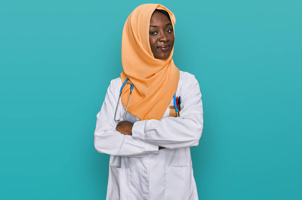 Hermosa mujer joven africana con uniforme de médico y cara feliz hijab sonriendo con los brazos cruzados mirando a la cámara. persona positiva.  - Foto, imagen