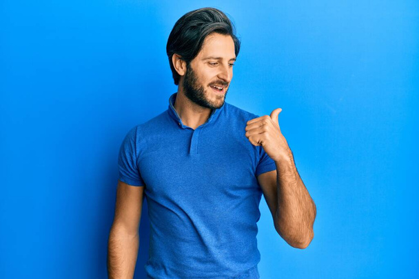 jonge latino man dragen casual blauw t shirt duim wijzen naar de zijkant glimlachen blij met open mond  - Foto, afbeelding