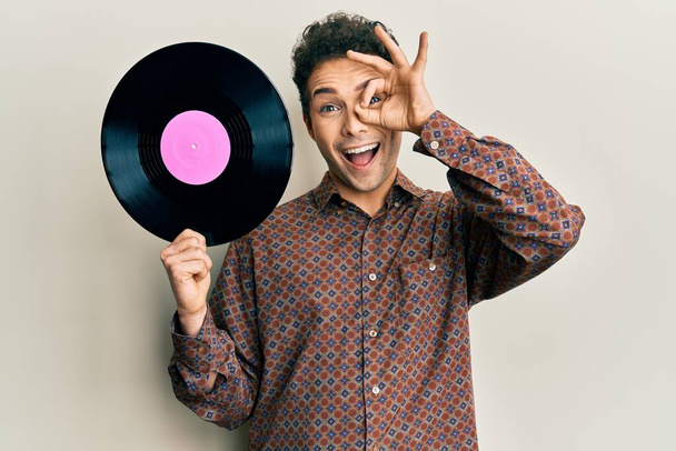Young handsome man holding vinyl disc smiling happy doing ok sign with hand on eye looking through fingers  - Foto, Imagen