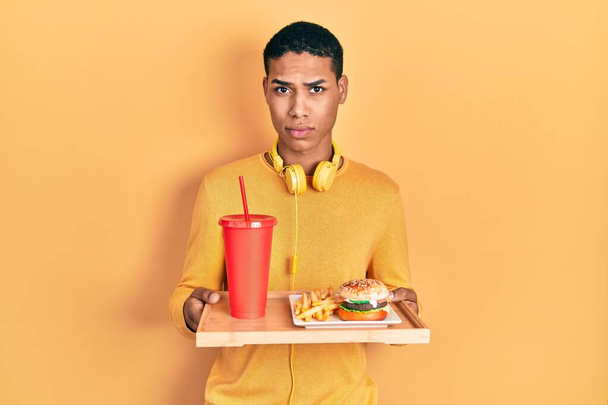 Young african american guy eating a tasty classic burger skeptic and nervous, frowning upset because of problem. negative person.  - Valokuva, kuva