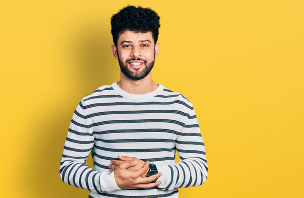 Der junge arabische Mann mit Bart trägt einen lässig gestreiften Pullover mit zusammengelegten Händen und Daumen, der entspannt und fröhlich lächelt. Erfolg und Optimismus  - Foto, Bild