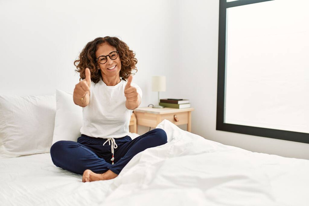 Middle age hispanic woman sitting on the bed at home approving doing positive gesture with hand, thumbs up smiling and happy for success. winner gesture.  - Photo, Image