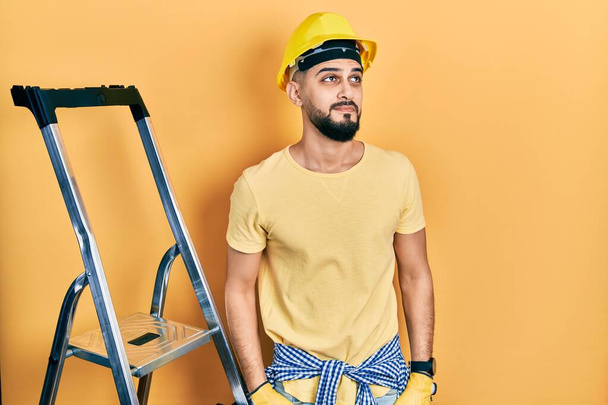 Handsome man with beard by construction stairs wearing hardhat smiling looking to the side and staring away thinking.  - Photo, Image
