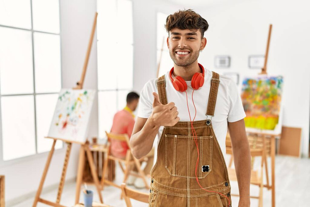 Молодий іспаномовний чоловік в художній студії робить щасливі великі пальці вгору жестом рукою. затверджуючий вираз, який дивиться на камеру, що демонструє успіх
.  - Фото, зображення