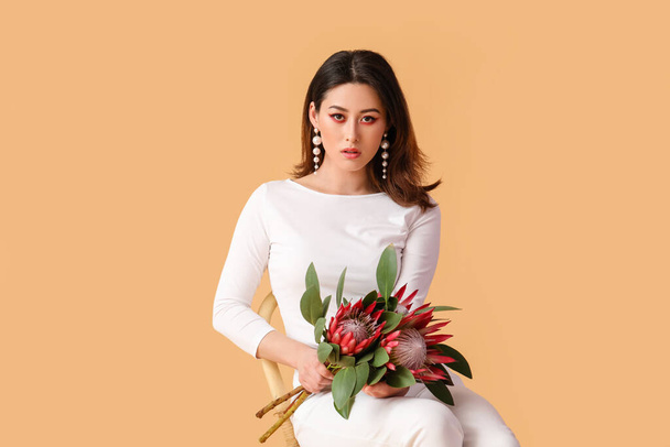 Beautiful young woman with protea flowers sitting on chair against color background - Foto, afbeelding