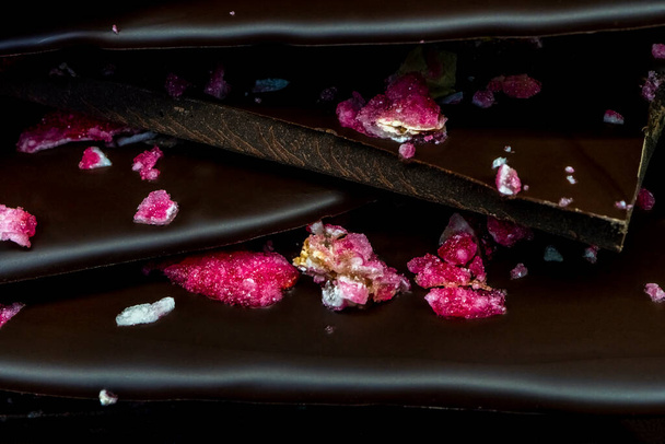 Πλακάκια μαύρης σοκολάτας με κόκκινα αποξηραμένα και φρέσκα μούρα και ζαχαρωμένα φύλλα λουλουδιών. Μαύρο φόντο, κοντινό. - Φωτογραφία, εικόνα