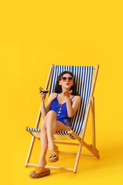Beautiful young woman with cocktail blowing kiss while sitting on deck chair against color background - Φωτογραφία, εικόνα