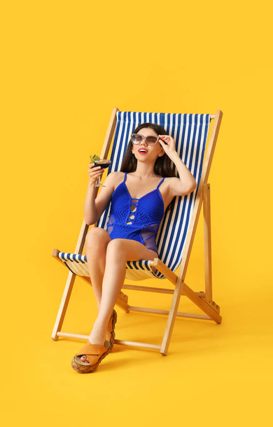 Beautiful young woman with cocktail sitting on deck chair against color background - Foto, afbeelding