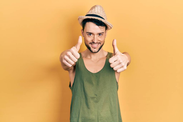Jeune homme hispanique portant un chapeau d'été approuvant faire un geste positif avec la main, le pouce levé souriant et heureux pour le succès. geste gagnant.  - Photo, image