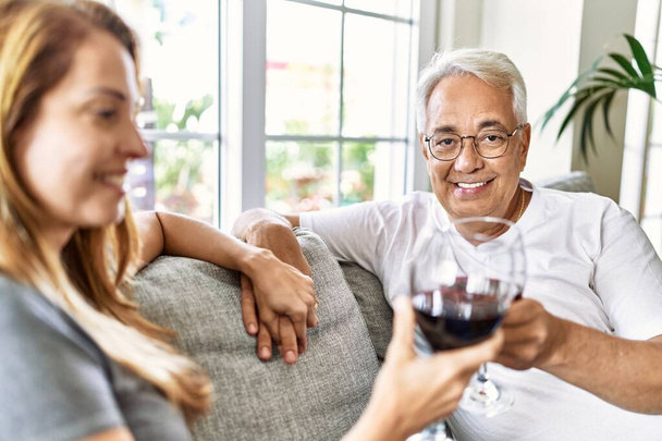 Μεσήλικες ισπανόφωνοι ζευγάρι χαμογελάνε ευτυχισμένοι πίνοντας κρασί καθισμένοι στον καναπέ στο σπίτι. - Φωτογραφία, εικόνα