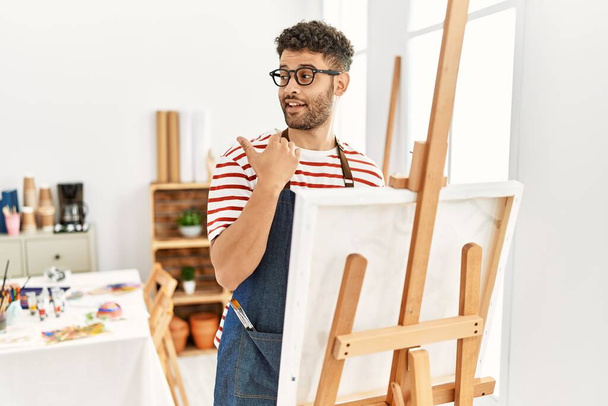 Arabi nuori mies art studio hymyilee onnellinen kasvot etsivät ja osoittaa sivuun peukalo ylöspäin.  - Valokuva, kuva