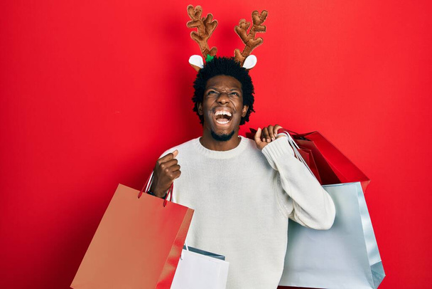 Joven hombre afroamericano con sombrero de navidad de ciervo sosteniendo bolsas de compras enojado y loco gritando frustrado y furioso, gritando con ira mirando hacia arriba.  - Foto, Imagen