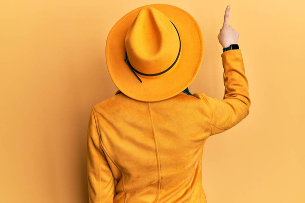 Young modern girl wearing yellow hat and leather jacket posing backwards pointing ahead with finger hand  - Fotoğraf, Görsel